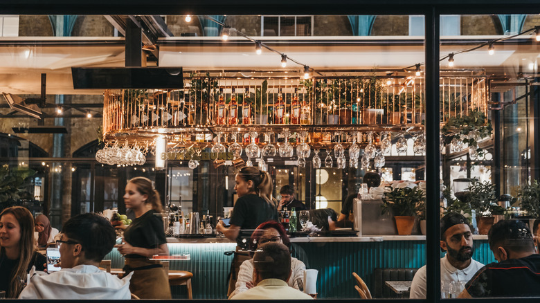 Daytime interior of a busy restaurant with wine glasses hanging above bar