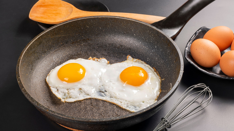 Two fried eggs in a pan next to some uncooked eggs and utensils