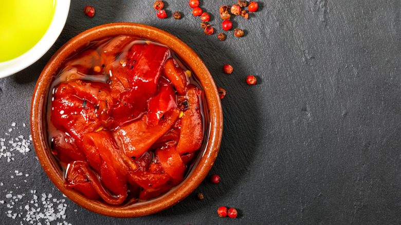 A bowl of roasted red peppers styled with olive oil and red peppercorns on a dark background
