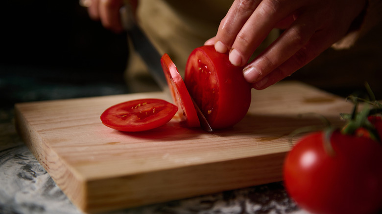 Someone slicing a fresh tomato on a wooden cutting board next to some whole tomatoes