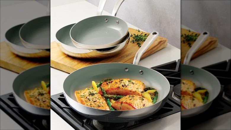 Three stainless steel pans, one on stovetop with salmon filets and two sitting on top of a cutting board.
