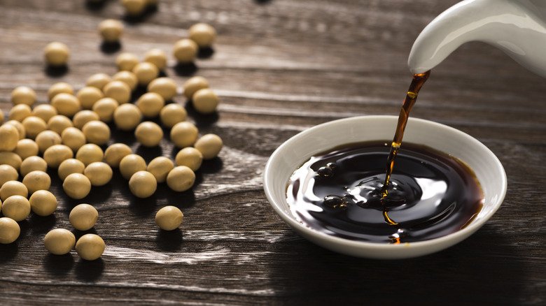 Soy sauce being poured into a white dish next to soybeans on a wooden background