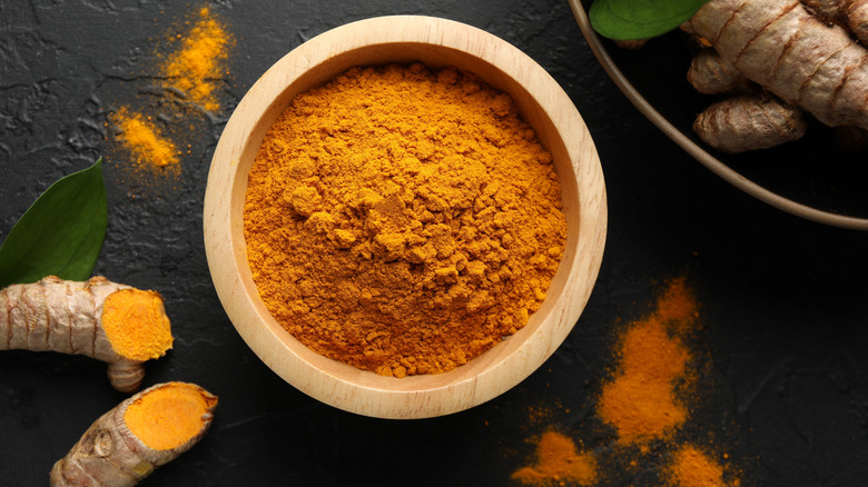 Wooden bowl of ground turmeric on black background with turmeric roots nearby