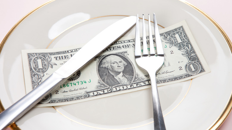 A dollar bill under a metal knife and fork on a gold-rimmed white plate