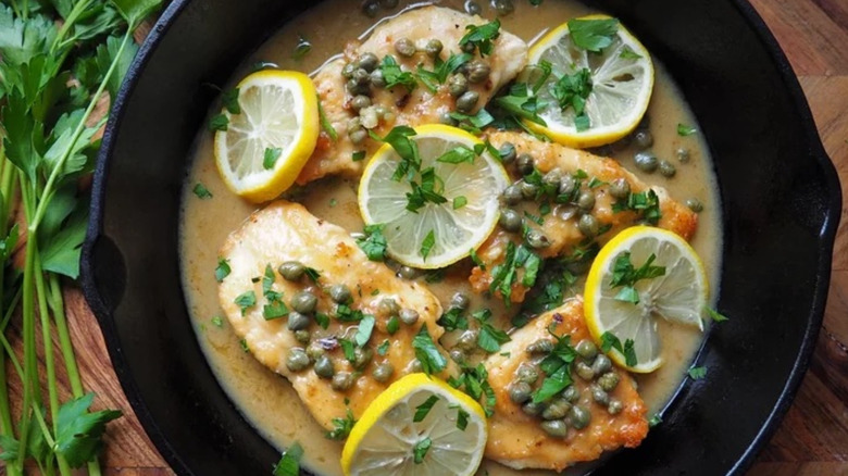 Skillet with chicken, caper sauce, lemon slices, and herbs.