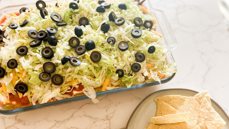 7-layer dip topped with shredded lettuce and black olives in a glass baking dish with tortilla chips off to the side