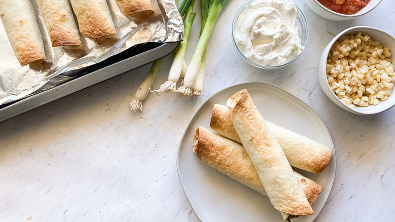 three taquitos stacked up on plate with ingredients like green onion, sour cream, corn, and salsa scattered around it