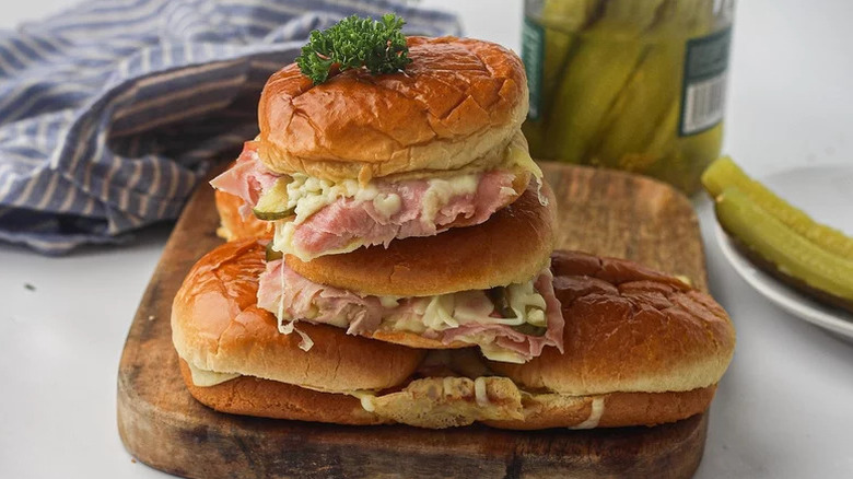 five ham and cheese sliders stacked on top of one another on wooden cutting board with lettuce garnish on top and pickle jar in background
