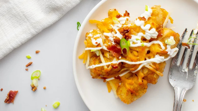 one serving of loaded baked potato totchos on a white plate with a fork to the side. Totchos are tater tots topped with cheese and sour cream.