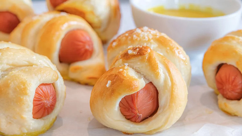 close up of several mini pretzel hot dogs sprinkled with salt with a bowl of cheese sauce in the background