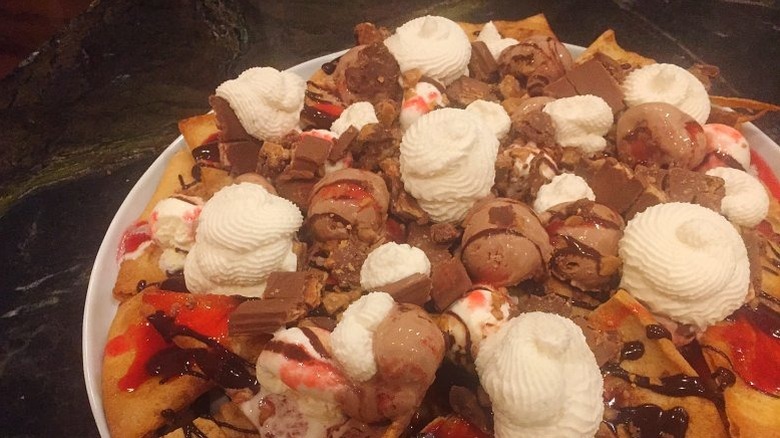 close up of dessert nachos on a plate, pita chips drizzled with chocolate and raspberry syrups, topped off with scoops of chocolate ice cream, candy pieces, and whipped cream