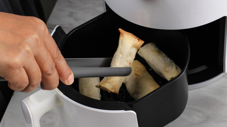 A hand holding tongs picking up one spring roll from inside an air fryer