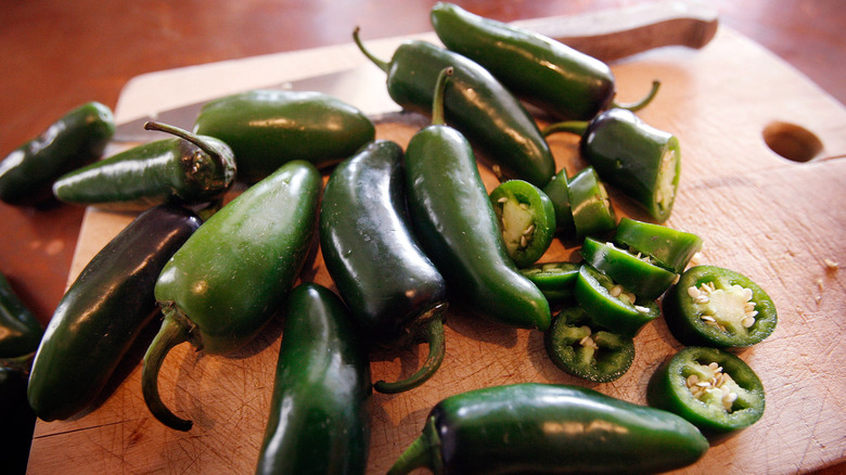 Multiple jalapeño peppers, some cut, on a wooden cutting board with a knife