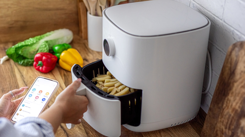 Person checking food cooking inside an air fryer while holding mobile device.