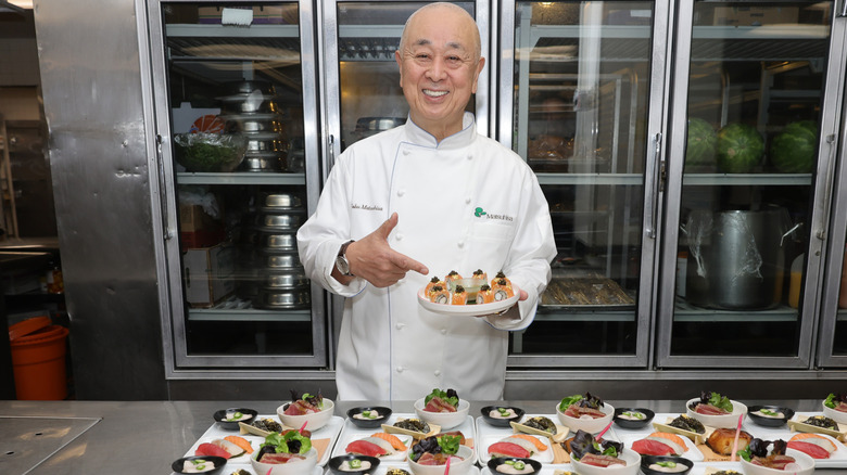 Chef Nobu Matushisa smiles and points to sushi rolls on a white plate inside a commercial kitchen