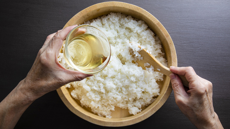 Hands holding sushi vinegar in a glass bowl and wooden paddle above white rice in a wooden bowl