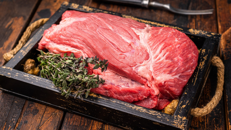 Piece of raw beef top round on a rustic tray with rope handles and sprigs of herbs.