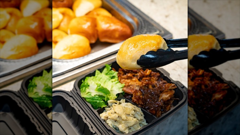 Person at buffet restaurant taking dinner roll from tray with tongs.