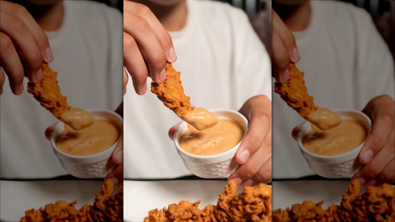 Person dunking chicken tender into a cup of creamy orange sauce.