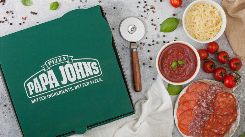 Green Papa John's pizza box next to pizza cutter, sauce, and other pizza ingredients.