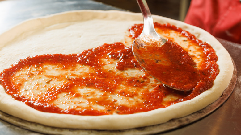 Large metal spoon spreading red sauce onto raw pizza dough.