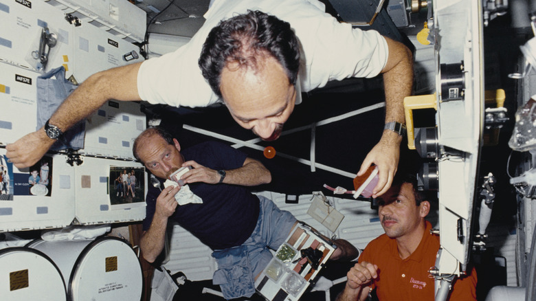 Floating astronaut attempting to drink a bubble of liquid while two other astronauts look on.