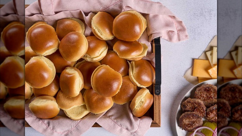 A tray piled high with brioche slider buns next to hamburger patties and toppings.