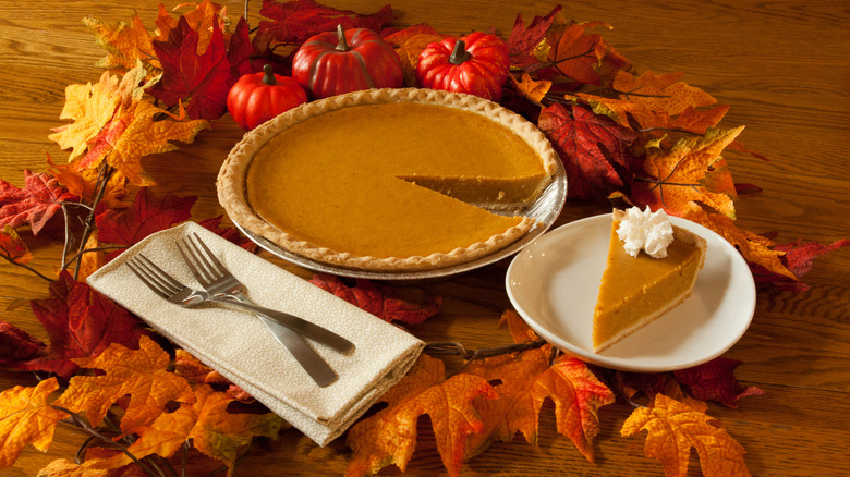 Pumpkin pie with a slice next to it on a plate amid festive fall leaves and mini pumpkins
