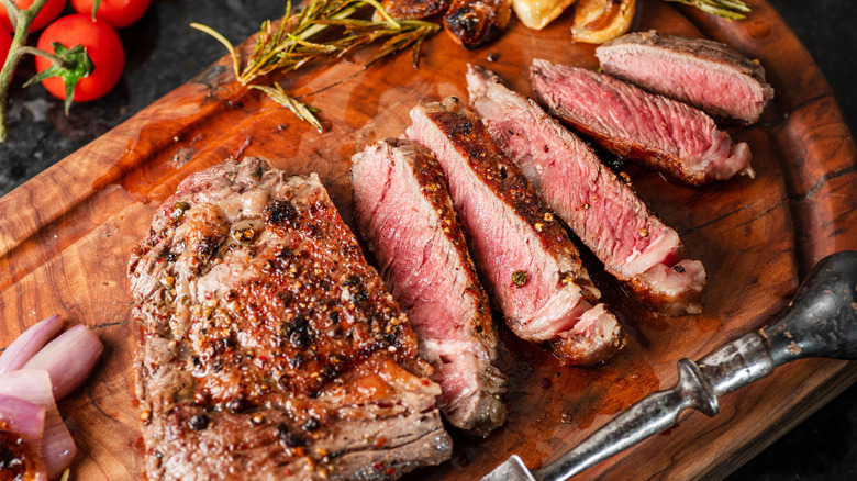 Ribeye steak cooked medium rare on a cutting board, sliced and next to metal serving fork and veggies.