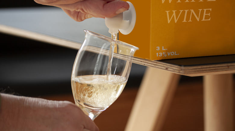 Hands dispensing white wine from yellow box on black and tan table into plastic glass