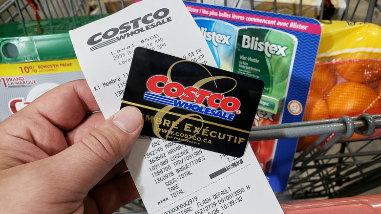 Person holding Costco card and receipt over a cart with groceries