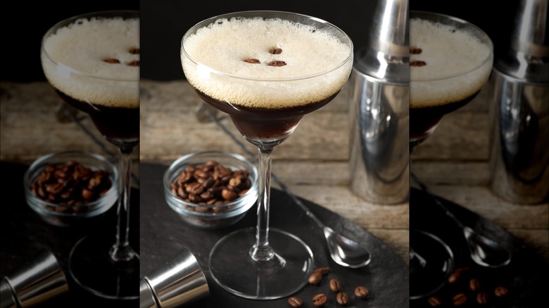 An espresso martini surrounded by a drink shaker, spoon, and coffee beans.