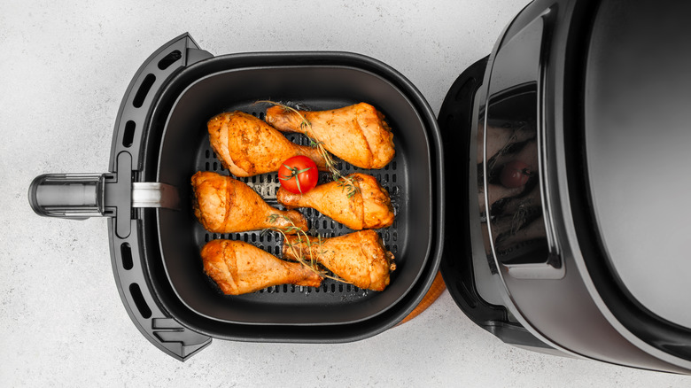 Overhead view of chicken drumsticks with a tomato and herbs in black basket of an air fryer