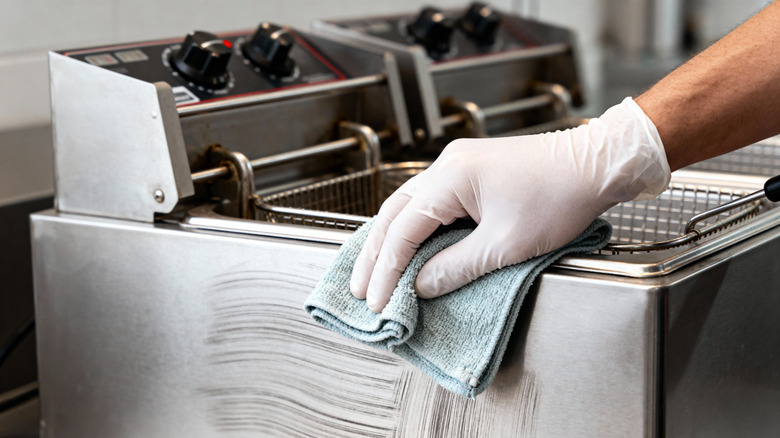 A person wearing a white glove uses a cloth to clean the outside of a deep fryer