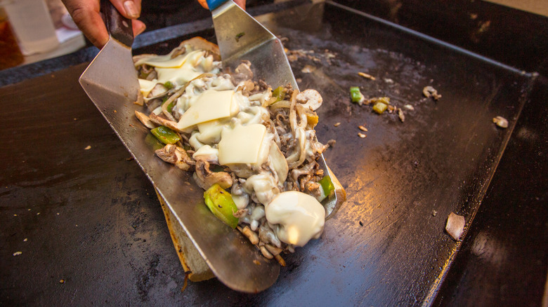 Person grilling a cheesesteak with meat, cheese, and vegetables between two spatulas.
