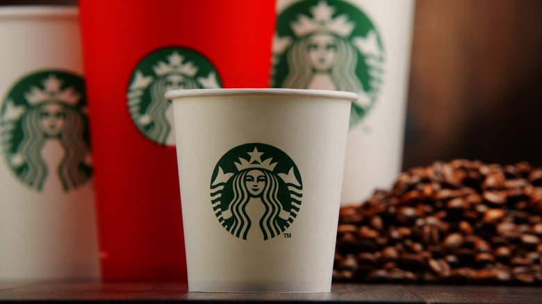 Various sizes of Starbucks cups, most white, one red, with coffee beans on wood table against brown wall