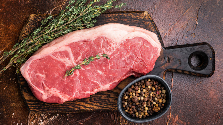 A piece of raw sirloin steak surrounded by herbs and peppercorns.