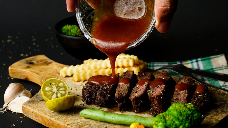 Hand pouring cup of steak sauce over sliced steak on cutting board