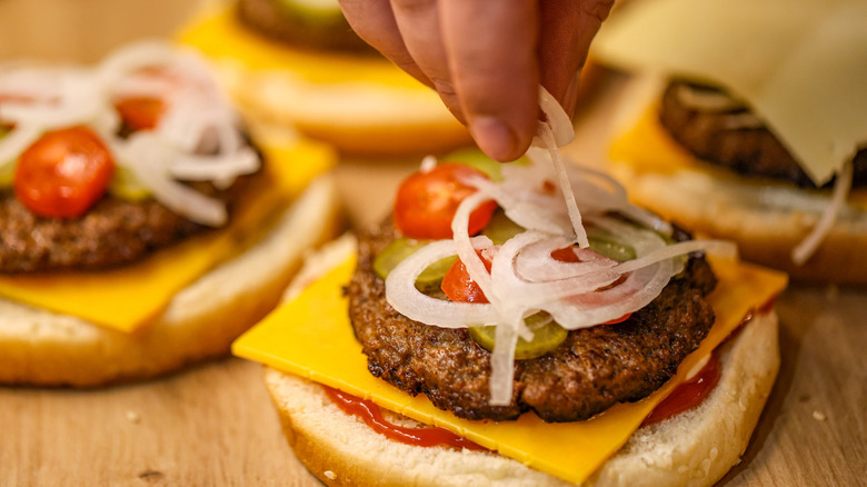 Assembling burgers on buns with cheese, onions, tomatoes and pickles, plus condiments.