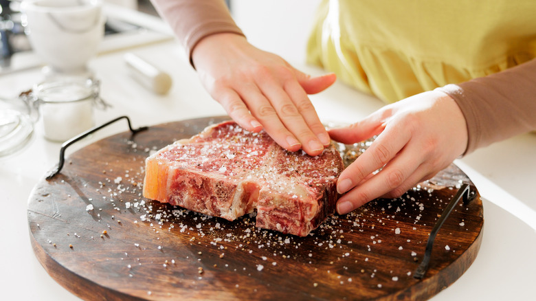 Person adding large crystals of salt to the outside of a raw steak on a wooden cutting board