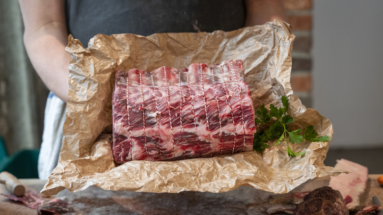 Person holding uncooked beef roast tied with string in waxy brown paper.