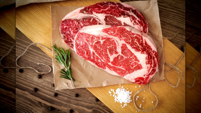 Two well-marbled ribeye steaks on wax paper on top of cutting board.