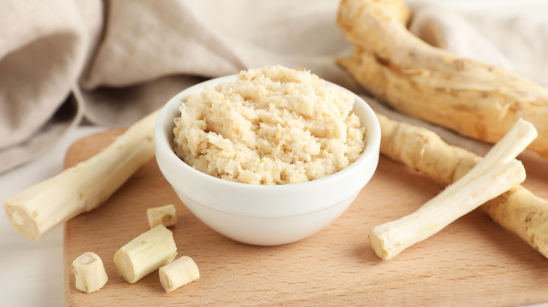 Freshly grated horseradish in white bowl surrounded by whole roots.