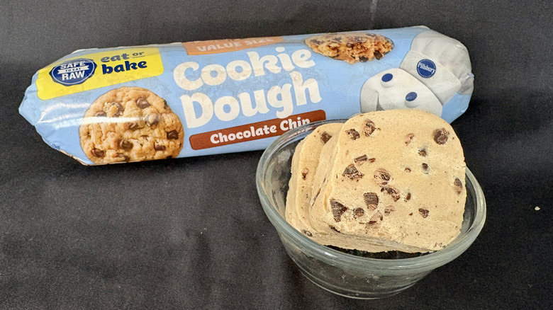 A log of Pillsbury cookie dough behind a glass bowl of slices of cookie dough