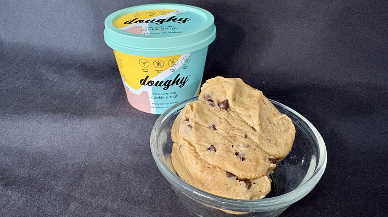 A scoop of chocolate chip cookie dough in a glass bowl with a Doughy branded tub behind