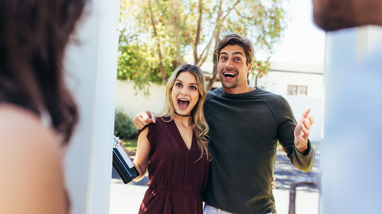 Two smiling guests at door, one holding bottle of wine
