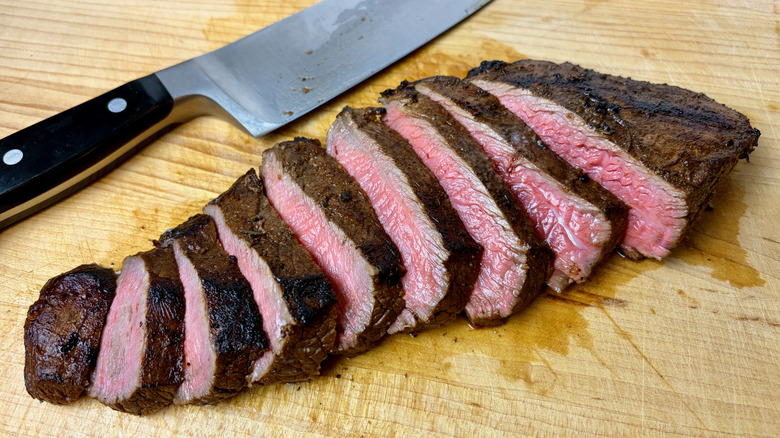 A cooked Trader Joe's Beef Flank Steak sliced on wooden cutting board next to knife.