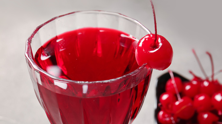 Closeup of a cocktail glass filled with a red beverage and one maraschino cherry, a plate of red cherries visible in the bottom corner