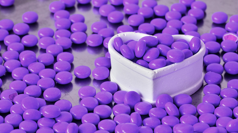 A white, heart-shaped container filled with violet candies, with more scattered on the table