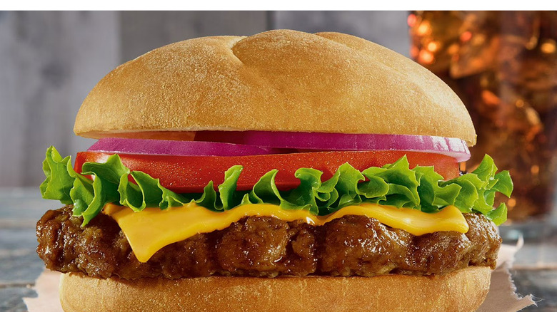 Sam's Club's cooked Angus cheeseburger on a napkin on a wooden table with a Coca-Cola or similar soft drink in the background in a glass cup